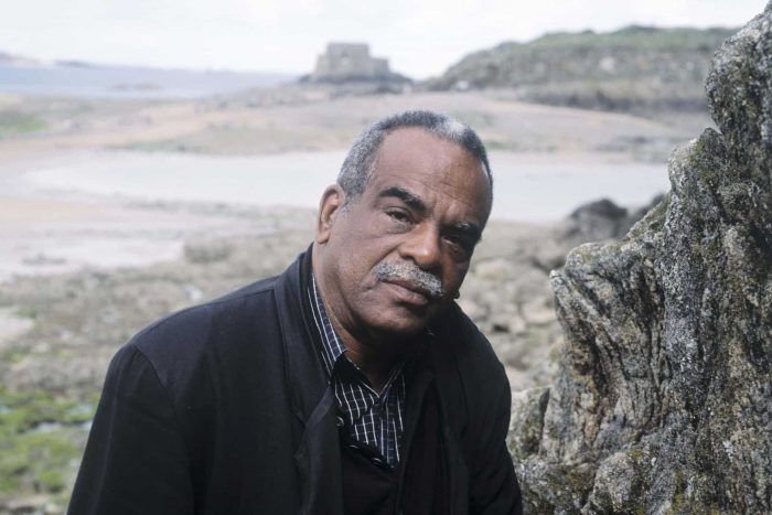 Fotografía de un hombre negro en una playa rocosa, con un bigote y pelo canosos